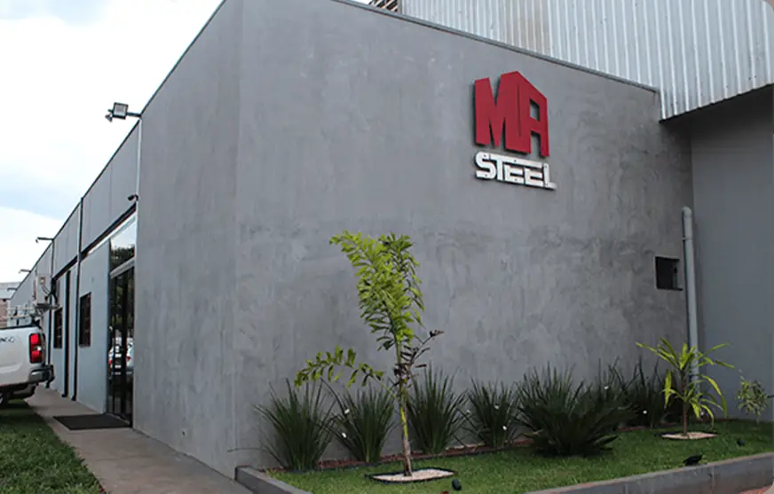 Fachada da MA Steel: galpão industrial cinza com o logotipo vermelho da empresa na parede e jardim na frente