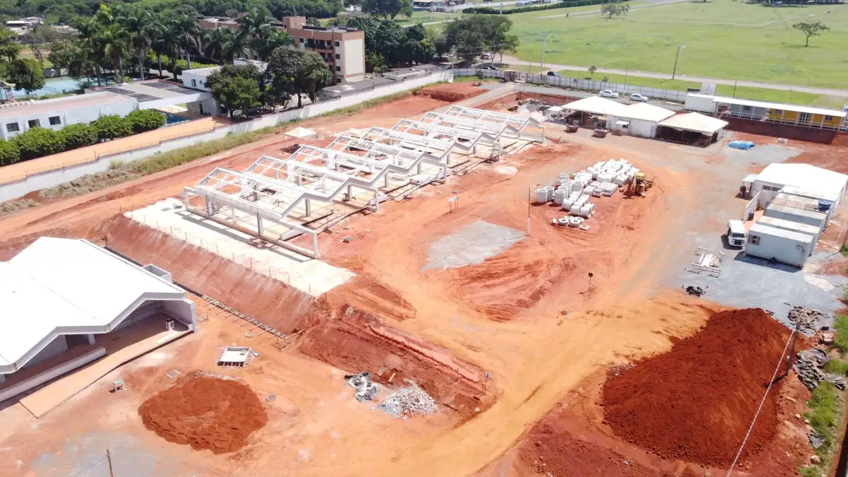 Vista aérea do canteiro mostrando os pórticos metálicos alinhados, caminhões e material estocado ao redor