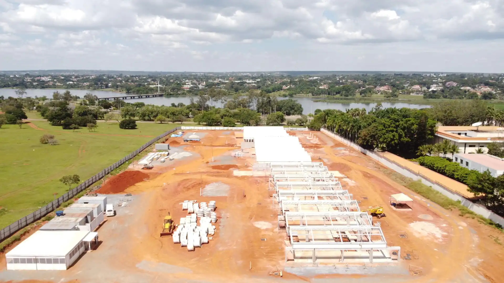 Vista aérea do canteiro de uma obra da MA Steel: galpões de estrutura metálica em montagem sobre terra vermelha, com peças pré-fabricadas empilhadas ao lado, e um lago e a cidade ao fundo